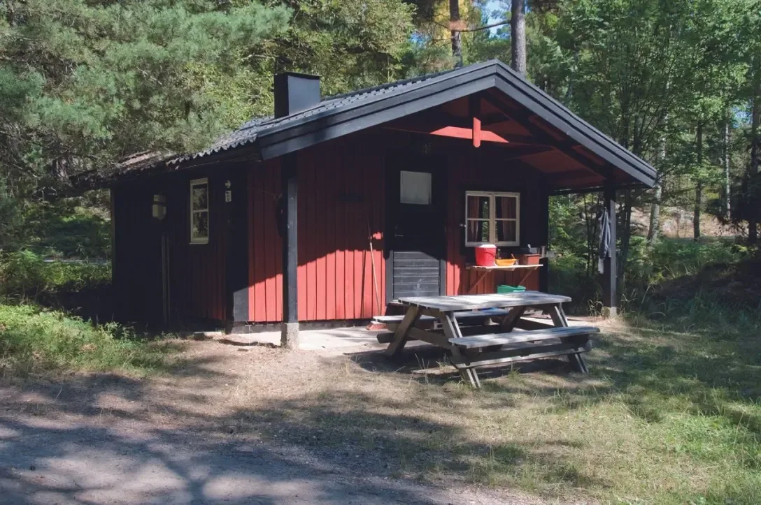 En röd stuga med svart tak och picknickbord står bland tallarna på Grinda.
