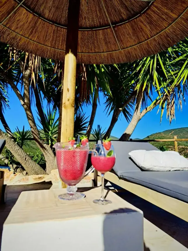 Två rosa fruktdrinkar står på ett bord framför en solsäng under ett halmtak, med palmer och berg i bakgrunden.