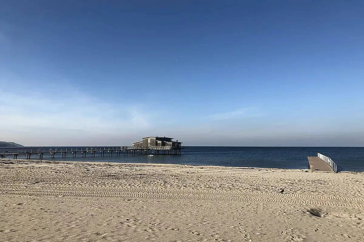 En bred sandstrand vid havet med en lång brygga och en byggnad längst ut under en klarblå himmel.