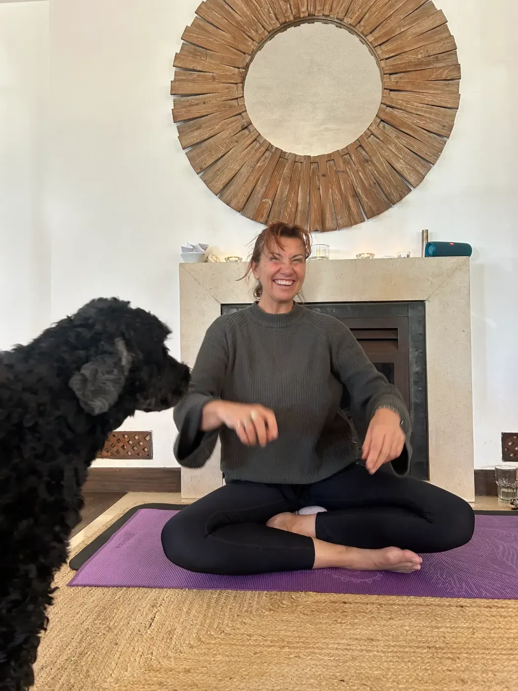 En leende person sitter i korslagd ställning på en lila yogamatta framför en öppen spis, medan en svart hund syns i förgrunden.