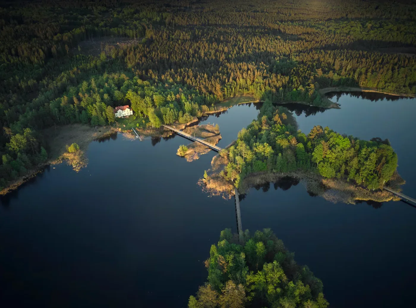 Flygvy över en skogsklädd sjö med en vit villa, brygga och broar mellan små öar.