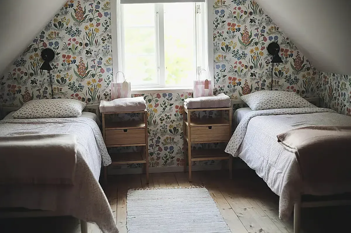 Ljust sovrum med två bäddade enkelsängar, träbord och färgglada blommiga tapeter under ett snedtak.