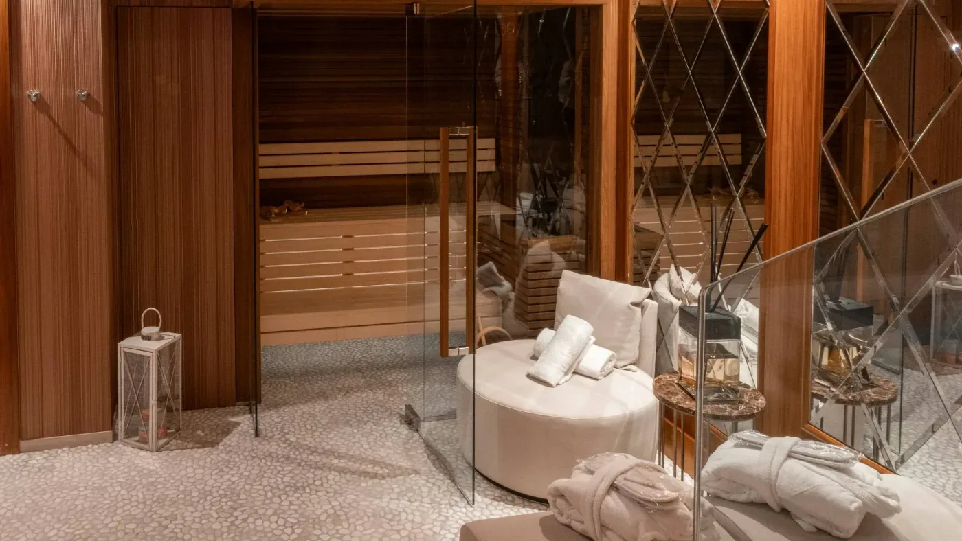 Ett varmt inrett spa med träpaneler, glasväggar, bastu, vita sittmöbler och hopvikta badrockar.