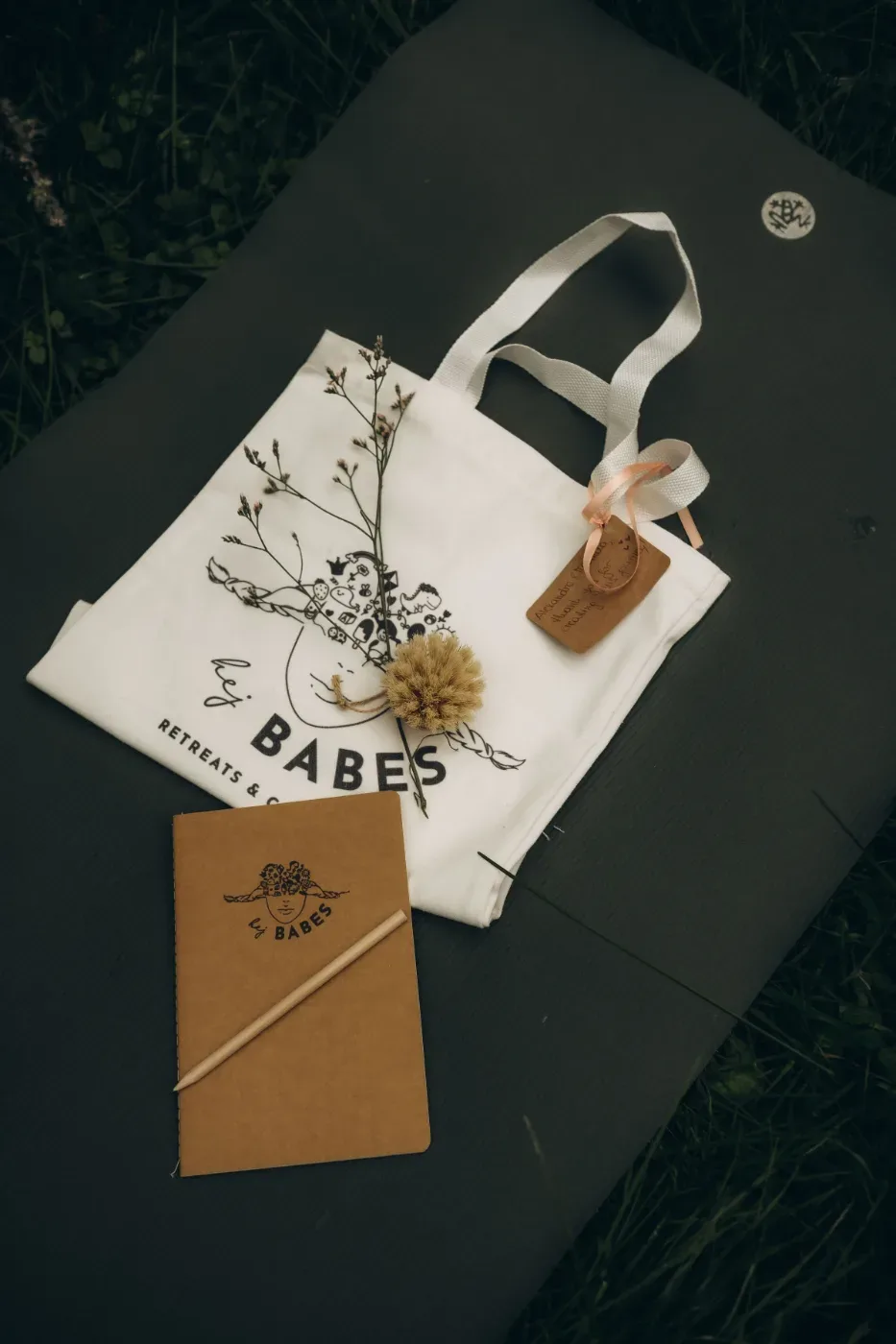 En tygkasse med trycket ”Babas Retreats &...” ligger på en mörk yta tillsammans med ett brunt anteckningsblock, en penna, torkade blommor och ett presentkort med band.