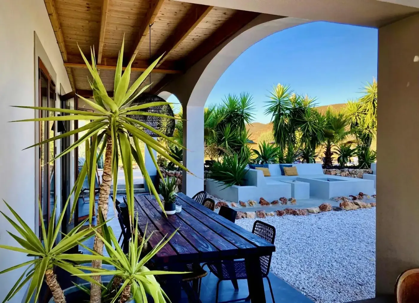 Skuggad uteplats med träbord och stolar, omgiven av palmliknande växter och vita sittbänkar mot ett soligt bergslandskap i Algarve.