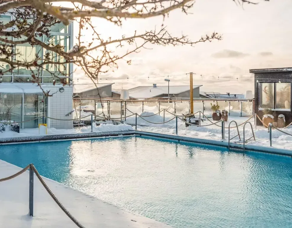 En uppvärmd utomhuspool omgiven av snö, byggnader och nakna träd i Torekov.