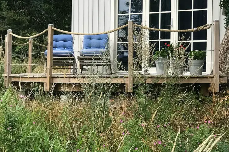 En veranda med blå sittdynor, krukväxter och repstaket framför ett vitt hus, omgiven av en blommande äng.