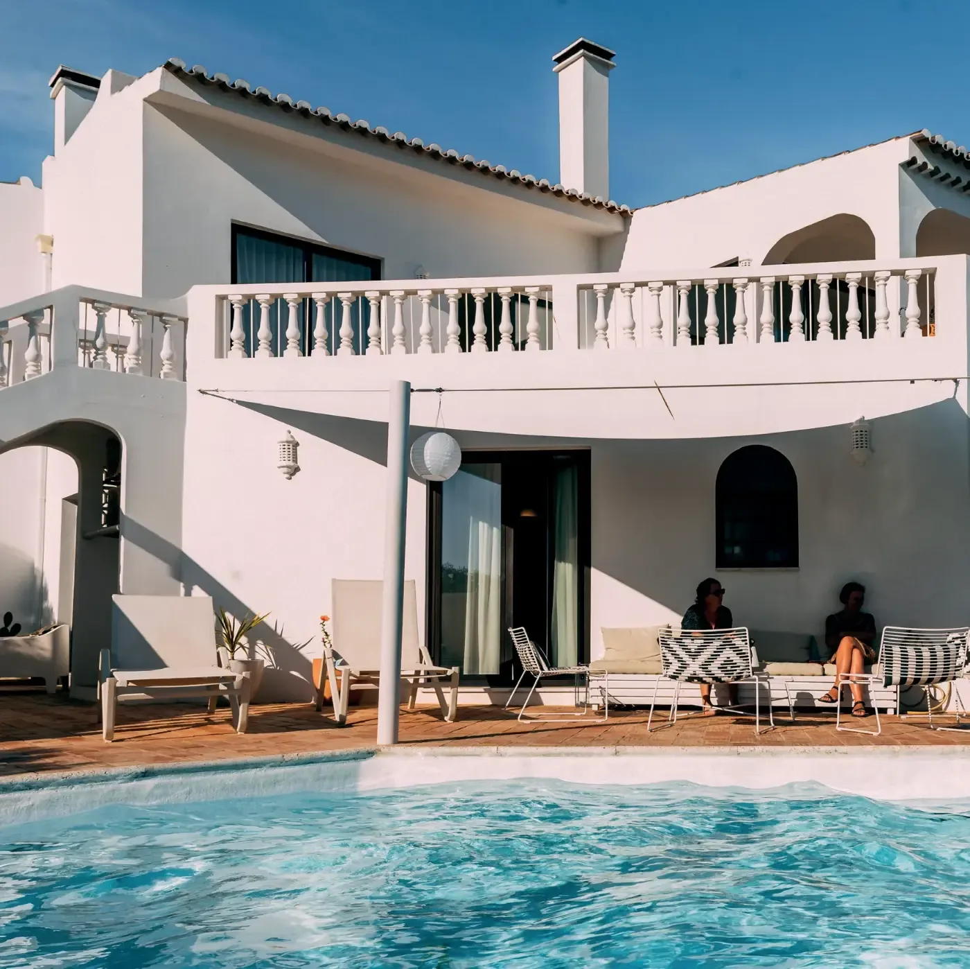 Vitkalkad villa med terrass, loungemöbler och två personer vid en pool under klarblå himmel i Algarve.