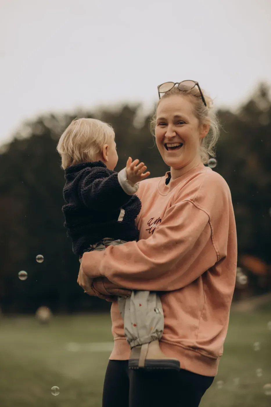 En leende vuxen håller ett litet barn i famnen utomhus, omgivna av flygande såpbubblor och grönska.