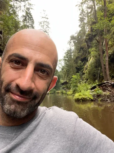 Yasser Dehnavi ler mot kameran framför en stilla bäck omgiven av frodig skog.