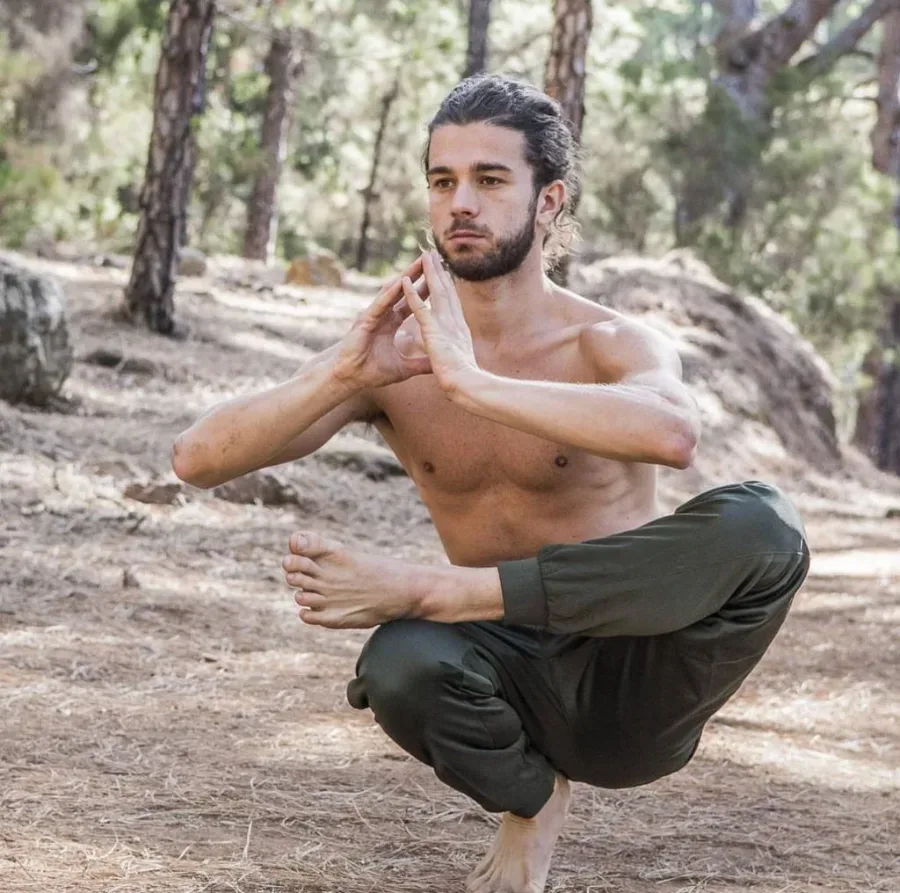 Barfotad man i mörka kläder gör en balanserande yogaposition i en tallskog.