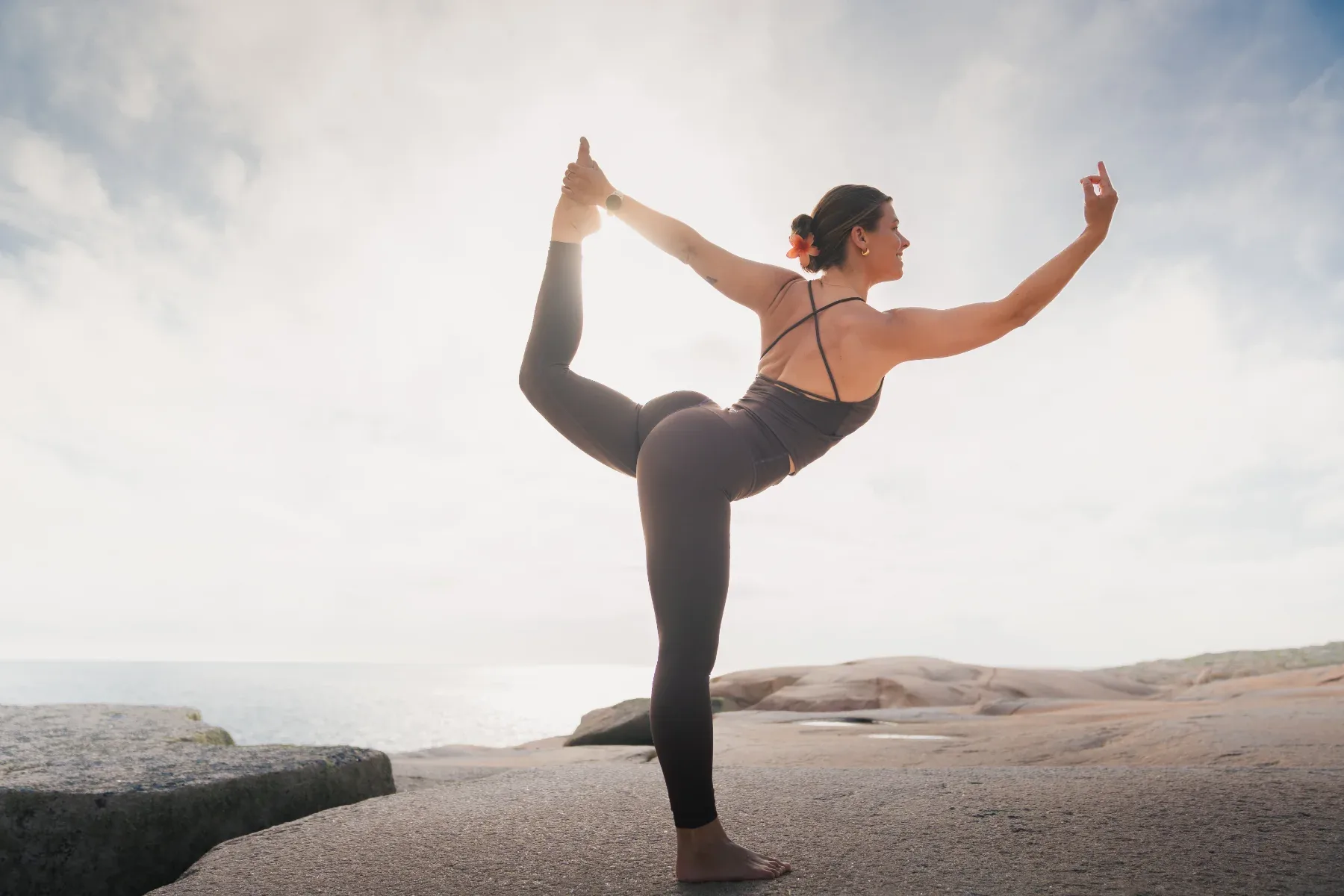 En kvinna i svart träningsklädsel gör en stående yogaposition på släta klippor vid havet.