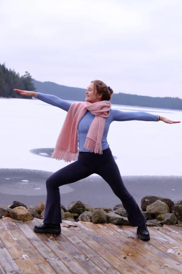 En person i rosa halsduk står i en yogaposition på en träbrygga vid en frusen sjö.