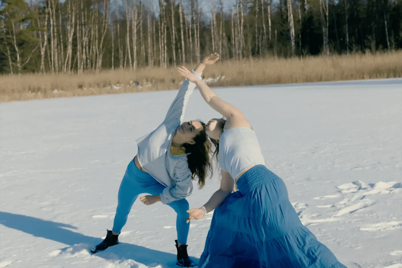 Två personer gör en yogaliknande rörelse tillsammans på en snötäckt sjö framför en björkskog.