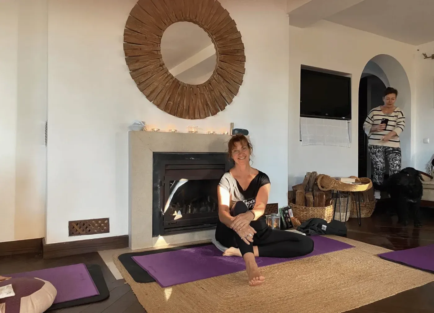 En kvinna sitter på en yogamatta framför en öppen spis, medan en annan person står i bakgrunden i ett ljust rum.