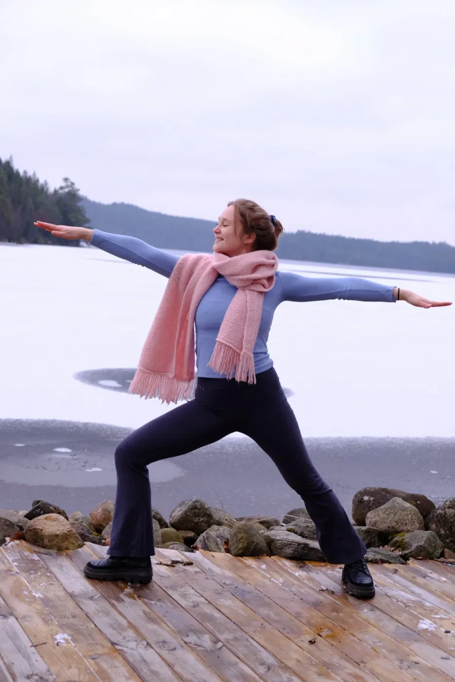 En kvinna i rosa halsduk och blå tröja står i en yogaposition på en träbrygga vid en frusen sjö, omgiven av skog och klippor.