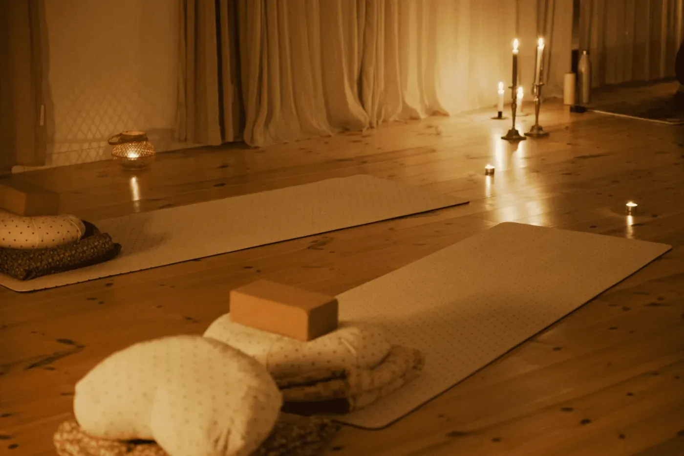Ett varmt upplyst rum med yogamattor, kuddar och block på trägolvet, omgivet av levande ljus.